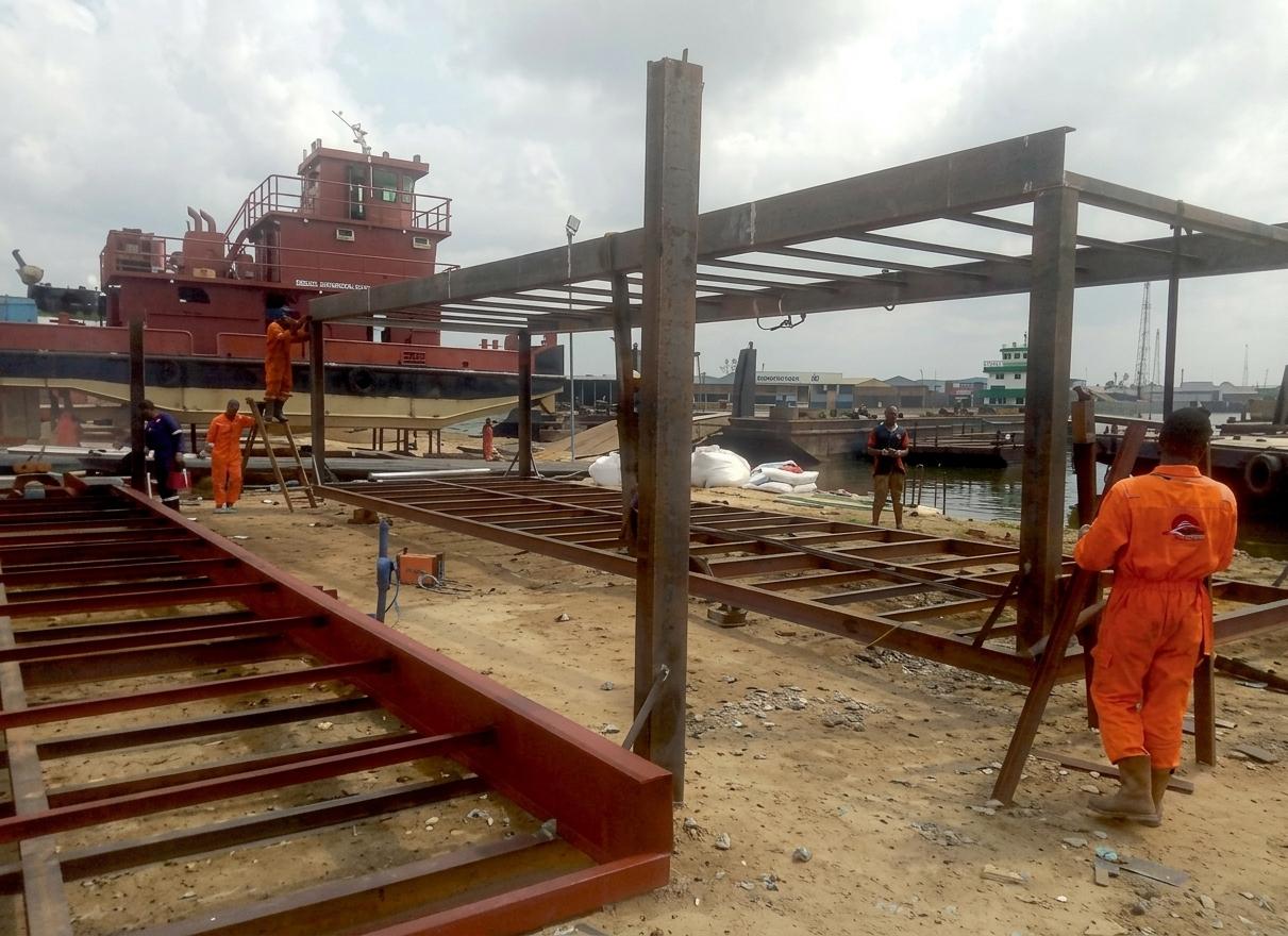 Crew in orange coveralls erecting a steel frame on a waterfront yard beside a tugboat
